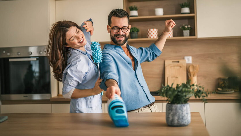 Zwei Personen stehen in einer Küche vor einer Arbeitsfläche. Eine Person hält einen Staubwedel, die andere ein Handgerät, während beide die Arme ausgestreckt haben.