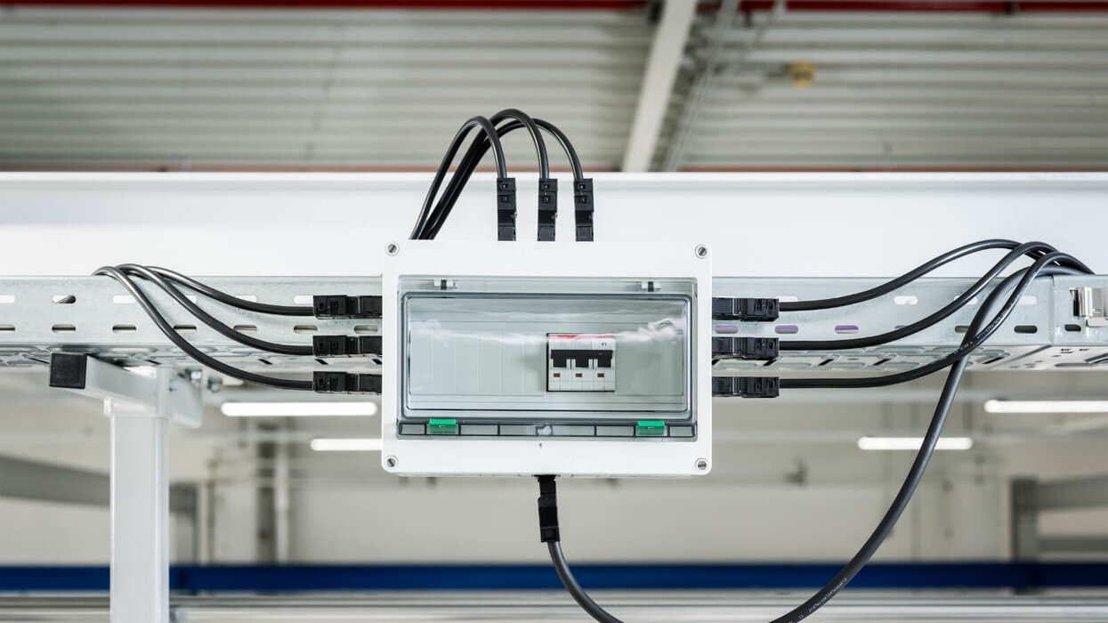 Das Bild zeigt ein montiertes Installationsgehäuse mit mehreren angeschlossenen Stromkabeln an einer Kabelrinne in einer Hallendecke.