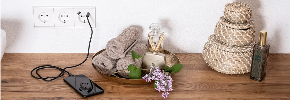 Das Bild zeigt ein Smartphone, das auf einer Holzablage über eine weiße Dreifach-Steckdose mit USB-Anschluss geladen wird. Daneben befinden sich eine Schale mit Handtüchern, ein Raumduft und Blumen.
