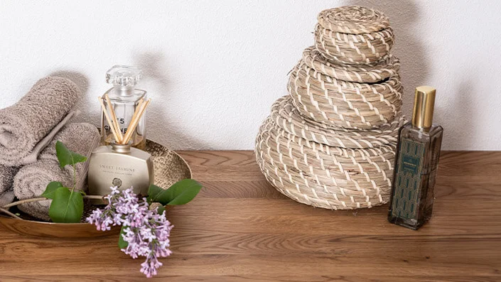 Das Bild zeigt ein Smartphone, das auf einer Holzablage über eine weiße Dreifach-Steckdose mit USB-Anschluss geladen wird. Daneben befinden sich eine Schale mit Handtüchern, ein Raumduft und Blumen.