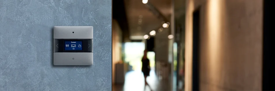 Das Bild zeigt ein Bedienelement des Herstellers Theben, welches an einer grauen Wand montiert ist. Im Hintergrund sieht man einen langen Flur.