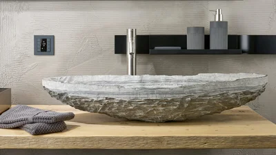 Ein Badezimmer mit einem einzigartigen Waschbecken aus Naturstein, das auf einer Holzplatte ruht. An der strukturierten Wand ist ein modernes Smart-Home-Bedienfeld angebracht. Über dem Waschbecken befindet sich eine schwarze Ablage mit einem Seifenspender und einem Zahnputzbecher. Graue Handtücher liegen auf der Holzplatte.