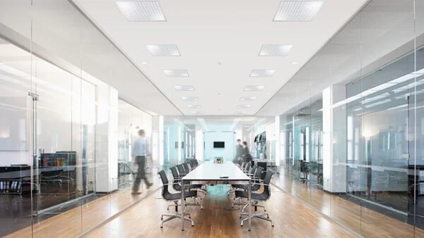 Das Bild zeigt einen Raum, in dessen Mitte ein großen, länglichen Konferenztisch mit modernen Stühlen an den Längsseiten. Rechts und links vom Tisch liegen Büros hinter Glaswänden. In der abgehängten Decke des Raumes sind in zwei parallel verlaufenden Reihen Leuchten angebracht. Einige Personen gehen durch den Raum. 