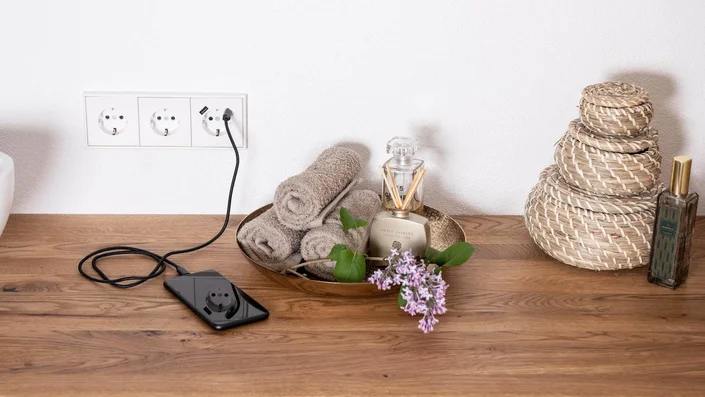 Das Bild zeigt ein Smartphone, das auf einer Holzablage über eine weiße Dreifach-Steckdose mit USB-Anschluss geladen wird. Daneben befinden sich eine Schale mit Handtüchern, ein Raumduft und Blumen.
