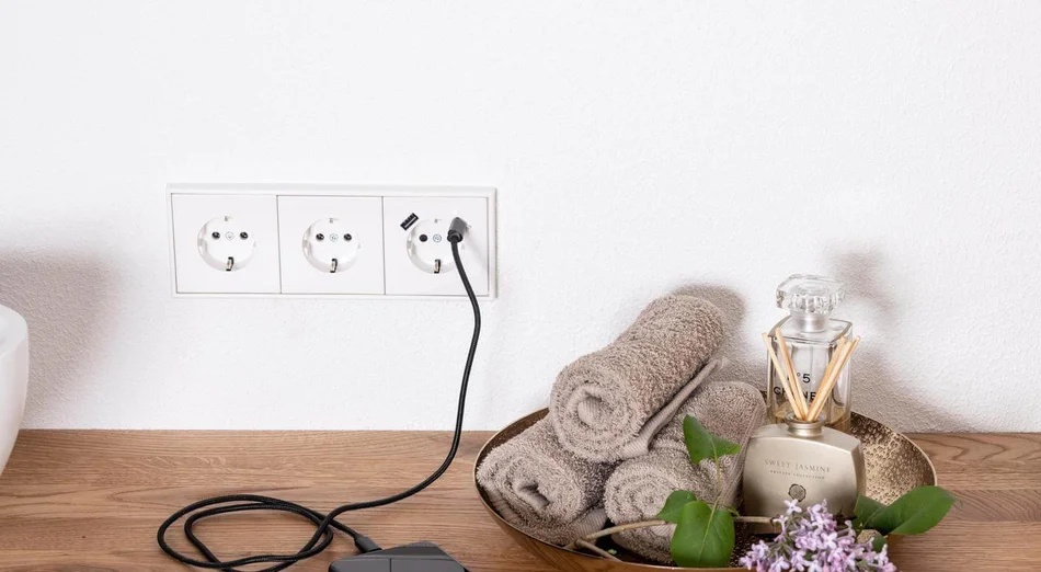 Das Bild zeigt ein Smartphone, das auf einer Holzablage über eine weiße Dreifach-Steckdose mit USB-Anschluss geladen wird. Daneben befinden sich eine Schale mit Handtüchern, ein Raumduft und Blumen.