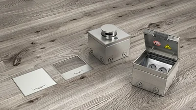 Verschiedene Bestandteile einer Bodensteckdose von OBO Bettermann auf dem braun-grauen Laminatboden liegend.