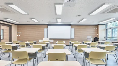 Das Bild zeigt ein helles, modernes Klassenzimmer mit vielen Tischen und Stühlen, einer großen Projektionsfläche vorn und mehreren rechteckigen Deckenleuchten.