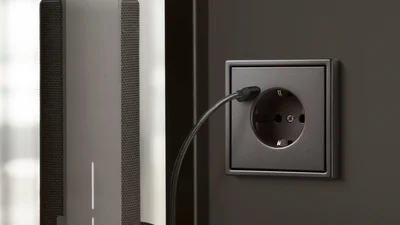 Das Bild zeigt eine moderne Steckdose in dunklem Design, in die ein Kabel eingesteckt ist. Daneben steht ein Lautsprecher.