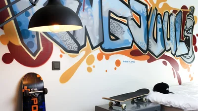 Das Bild zeigt ein modernes Jugendzimmer mit buntem Graffiti an der Wand. Zu sehen sind außerdem Skateboards, eine Hängelampe, ein Bett und ein Lichtschalter im urbanen Streetstyle-Design.