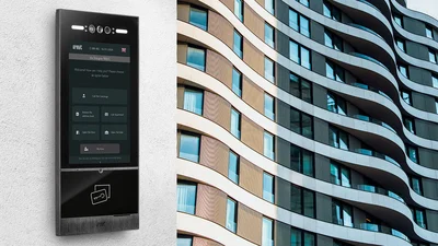 Moderne Türstation mit Touchdisplay an der Fassade eines mehrstöckigen Gebäudes mit geschwungener Architektur und großen Fensterflächen.
