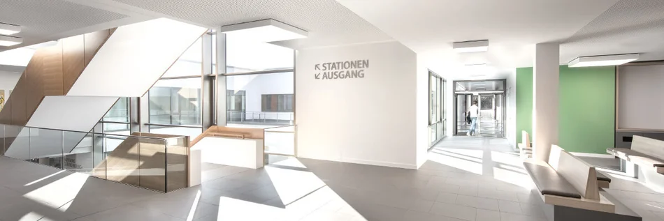 Das Bild zeigt einen modernen, hellen Eingangs- oder Wartebereich mit großen Fenstern, Sitzbänken und klarer Beschilderung für Stationen und Ausgang.