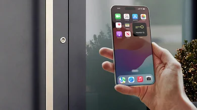 Eine Person hält ein Smartphone mit geöffneter App in der Hand, daneben ist ein Türschloss an einer Haustür zu sehen.