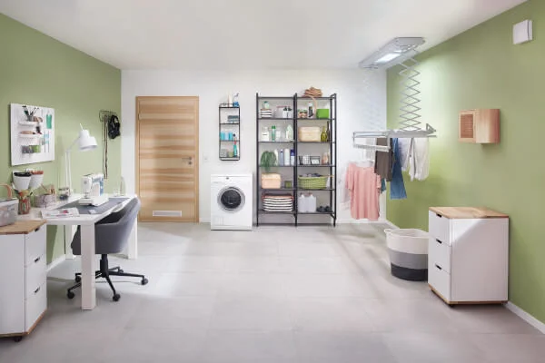 Das Bild zeigt einen Raum mit grünen Wänden, in dem ein Wäschetrockenständer und ein Regal mit verschiedenen Gegenständen stehen. Eine Waschmaschine befindet sich in der Ecke, und ein Schreibtisch mit einem Stuhl ist ebenfalls abgebildet.