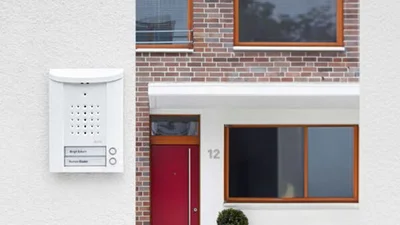 Bild der Ritto TwinBus Entravox Kompakt-Türstationan einer Hauswand. Im Hintergrund steht ein Haus mit roter Haustür