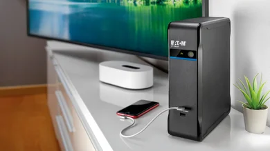 Das Bild zeigt ein weißes Sideboard auf dem eine schwarze 3P Ellipse USV steht, mit der ein Handy über ein Kabel verbunden ist. Links davon steht ein schwarzer Monitor Ein See mit umliegenden Bergen ist auf dem Bildschirm zu sehen. Rechts von der 3P Ellipse USV steht eine kleine Pflanze in einem weißen Übertopf. 