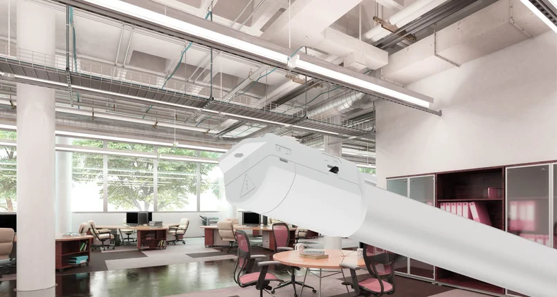 Das Bild zeigt ein modernes Großraumbüro mit mehreren Schreibtischen, Stühlen und Computern. Im Vordergrund ist eine vergrößerte Leuchte mit weißem Gehäuse zu sehen, die an der Decke montiert ist. An der Decke verlaufen außerdem sichtbare Kabeltrassen, Lüftungsrohre und weitere Installationen. Große Fenster lassen Tageslicht in den Raum.