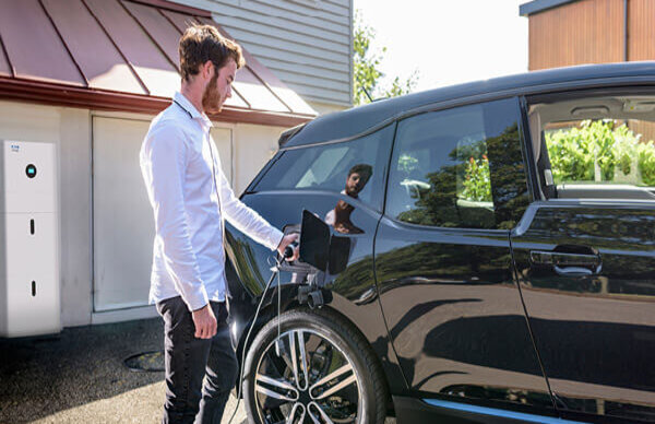 Das Bild zeigt einen Mann, der ein schwarzes Elektroauto im Außenbereich eines Hauses auflädt; im Hintergrund befindet sich eine weiße Geräteeinheit an der Hauswand.