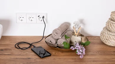 Das Bild zeigt ein Smartphone, das auf einer Holzablage über eine weiße Dreifach-Steckdose mit USB-Anschluss geladen wird. Daneben befinden sich eine Schale mit Handtüchern, ein Raumduft und Blumen.