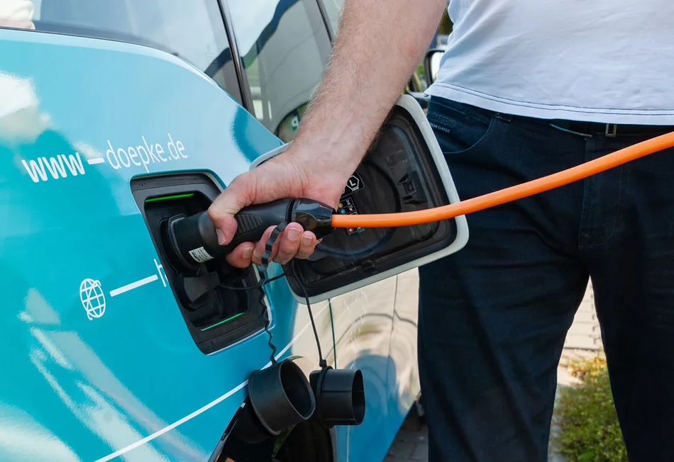 Das Bild zeigt eine Person, die ein orangefarbenes Ladekabel in die Ladebuchse eines türkisfarbenen Fahrzeugs steckt. Auf dem Fahrzeug steht die Internetadresse „www.doepke.de“.