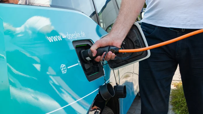 Doepke Fehlerstromschutzschalter für E-Mobility Das Bild zeigt eine Person, die ein orangefarbenes Ladekabel in die Ladebuchse eines türkisfarbenen Fahrzeugs steckt. Auf dem Fahrzeug steht die Internetadresse „www.doepke.de“.
