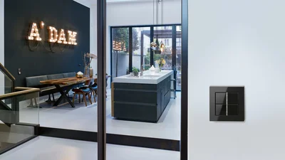 Eine moderne Wohnküche mit einem Essbereich, der eine dunkle Wand mit leuchtenden "A'DAM"-Buchstaben und einem Holztisch mit Sitzbank und Stühlen zeigt. Im Hintergrund ist eine Kücheninsel mit Spüle und Hängelampen sowie eine Glasfront zum Garten zu sehen. Rechts im Vordergrund befindet sich ein schwarzer Vierfach-Lichtschalter an einer weißen Wand.