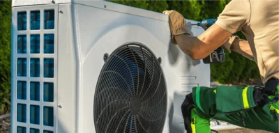 Heizung modernisieren. Eine Wärmepumpe ist zu sehen. Davor kniet ein Monteur.
