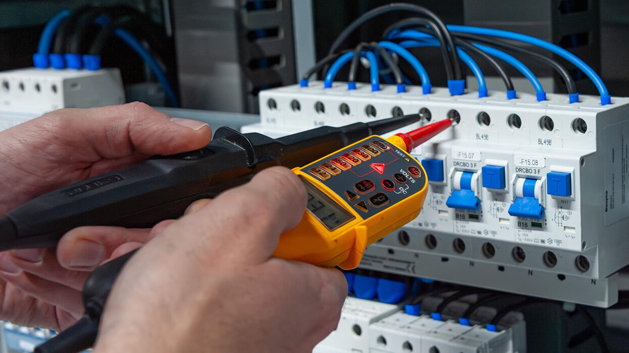 Das Bild zeigt eine Hand, die mit einem Spannungsprüfer elektrische Messungen an einem Schaltschrank durchführt. Mehrere blaue Kabel und Schutzschalter sind zu sehen.