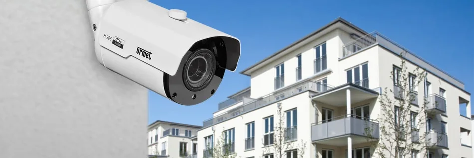 Das Bild zeigt eine Überwachungskamera von Urmet an einer Hauswand, mit Blick auf ein modernes Mehrfamilienhaus.