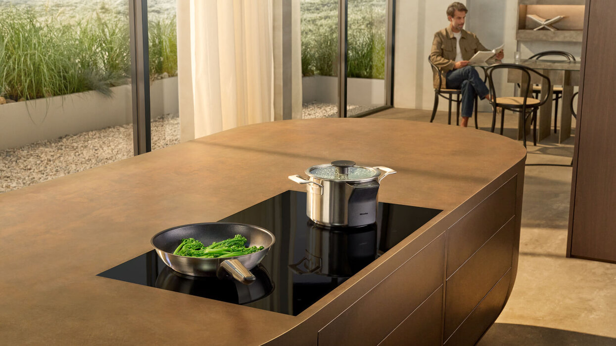 Das Bild zeigt eine moderne Kücheninsel mit schwarzem Kochfeld, einer Pfanne mit grünem Gemüse und einem Topf. Im Hintergrund sitzt ein Mann auf einem Stuhl und liest eine Zeitung.