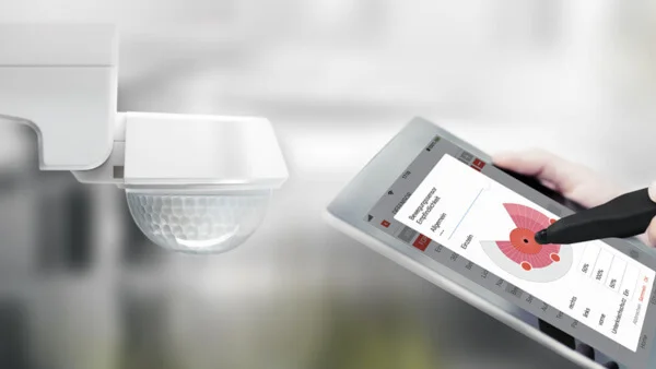 Das Bild zeigt auf der linken Bildseite einen weißen DEFENSOR Bewegungsmelder von ESYLUX. Auf der rechten Bildseite hält eine Hand einen schwarzen Laptop. Auf dem Display des Laptops eine Seite der ESY-App erkennbar. Eine andere Hand berührt mit dem ESY-Pen das Display des Tablets. Der Hintergrund des Bildes ist unscharf.