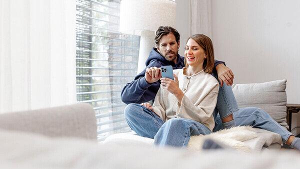 Das Bild zeigt ein entspanntes Paar, das gemeinsam auf einem Sofa sitzt. Sie halten ein Smartphone und schauen darauf, während im Hintergrund große Fenster mit Jalousien und eine helle, moderne Einrichtung zu sehen sind