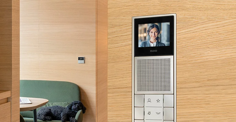 Innenstation zur Türkommunikation des Herstellers Jung direkt an der Holzwand montiert. Mit Display und verschiedenen Schaltern. Dahinter ein Wohnraum mit einem Sofa. 