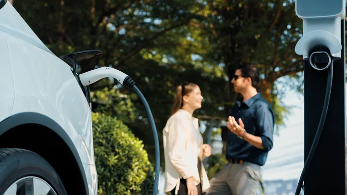 Das Bild zeigt ein E-Auto, welches gerade mit Strom betankt wird. Im Hintergrund sind zwei Personen zu sehen.