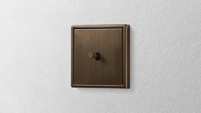 Das Bild zeigt einen messingfarbenen, rechteckigen Schalter mit Kipphebel an einer weißen Wand