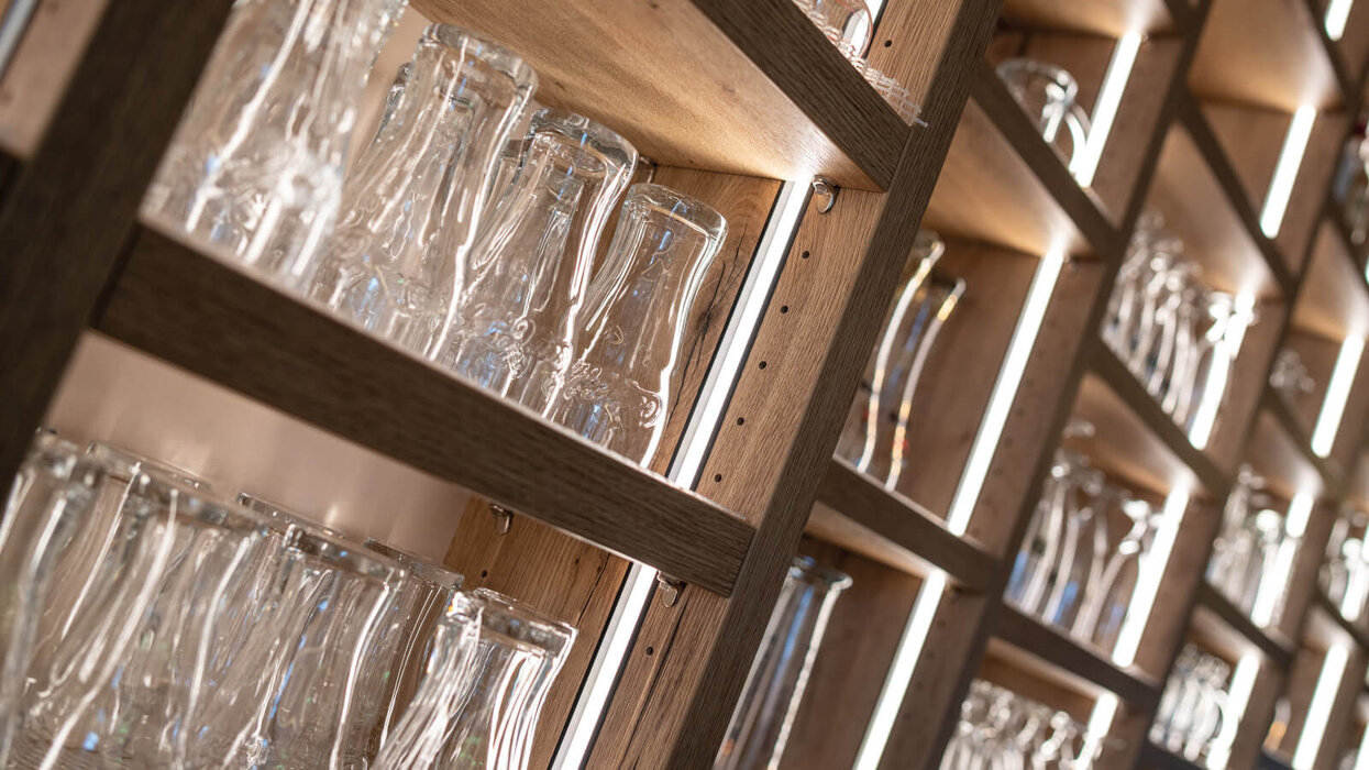 Zu sehen sind beleuchtete Holzregale mit vielen Gläsern in einer warmen, eleganten Innenraumgestaltung. Die Perspektive ist schräg und betont die gleichmäßige Regalbeleuchtung.
