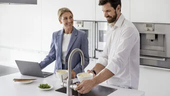Das Bild zeigt zwei Personen in einer modernen Küche. Eine Person bedient den Wasserhahn.