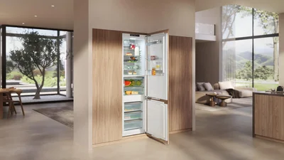 Das Bild zeigt eine moderne, offene Küche mit einem integrierten Einbau-Kühl-Gefriergerät. Die Türen des Kühlschranks sind geöffnet und geben den Blick auf übersichtlich eingeräumte Lebensmittel frei. Große Fenster und viel Tageslicht verleihen dem Raum eine helle, elegante Atmosphäre.