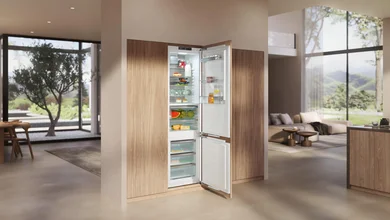 Das Bild zeigt eine moderne, offene Küche mit einem integrierten Einbau-Kühl-Gefriergerät. Die Türen des Kühlschranks sind geöffnet und geben den Blick auf übersichtlich eingeräumte Lebensmittel frei. Große Fenster und viel Tageslicht verleihen dem Raum eine helle, elegante Atmosphäre.