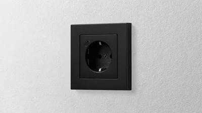 Das Bild zeigt eine Steckdose des Herstellers Jung in der Farbe Schwarz, die an einer Wand montiert ist.