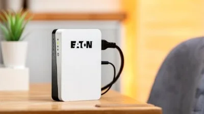 Der Bildfokus liegt bei einer Eaton 3S Mini USV, die auf einer Tischplatte aus Holz steht. An die Eaton 3S Mini USV sind zwei schwarze Kabel angeschlossen. Der Hintergrund ist verschwommen. Rechts ist die blaugraue Rückenlehne eines Stuhls erkennbar, links eine kleine Pflanze in weißem Übertopf..