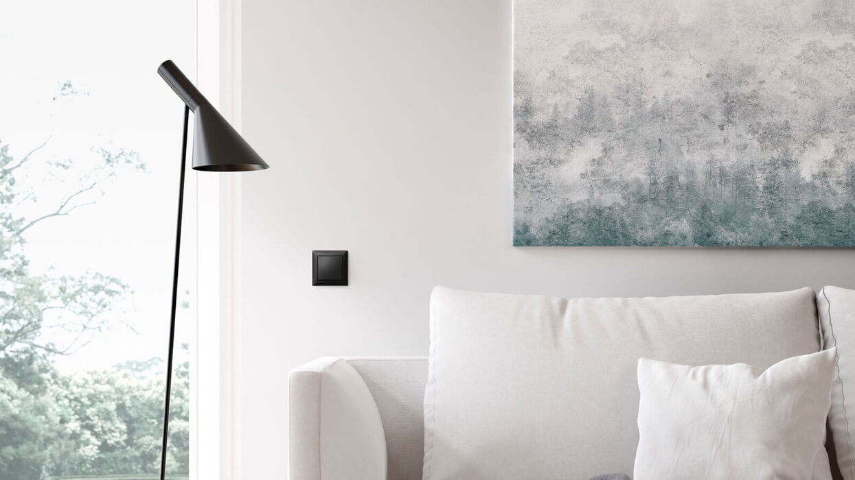 Ein minimalistisches Wohnzimmer mit hellem Sofa, schwarzer Stehleuchte und einem schwarzen Lichtschalter an der Wand, ergänzt durch ein abstraktes Wandbild und Tageslicht von außen.