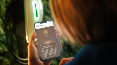 Das Bild zeigt eine Person, die ein Smartphone in der Hand hält. Auf dem Display ist eine Webseite mit dem Logo „MENNEKES“ zu sehen. Im Hintergrund leuchtet eine Wallbox.