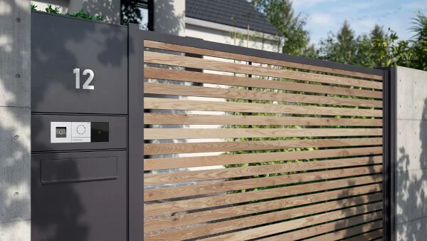 Das Bild zeigt einen Zaun mit großem Holztor. Links neben dem Tor befindet sich ein weißes Türkommunikationssystem mit schlüssellosem Zugang.