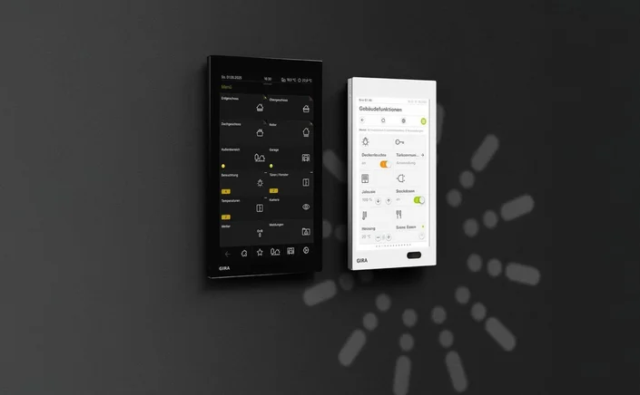 Das Bild zeigt zwei nebeneinander an einer Wand montierte Touch-Bedienpanels, eines in Schwarz und eines in Weiß, beide mit Benutzeroberflächen und dem GIRA-Logo. Auf den Displays sind verschiedene Symbole, Menüs und Steuerungsoptionen zu sehen.