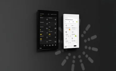 Das Bild zeigt zwei nebeneinander an einer Wand montierte Touch-Bedienpanels, eines in Schwarz und eines in Weiß, beide mit Benutzeroberflächen und dem GIRA-Logo. Auf den Displays sind verschiedene Symbole, Menüs und Steuerungsoptionen zu sehen.