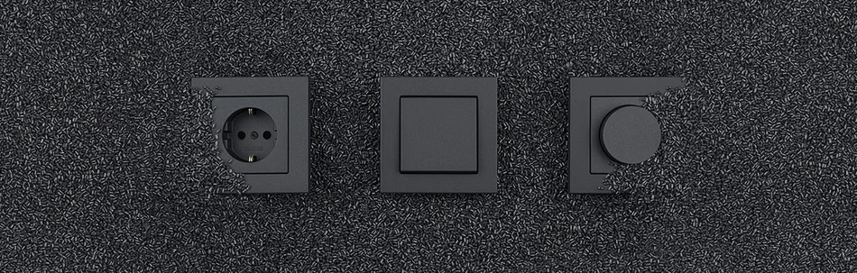 Das Bild zeigt eine schwarze Steckdose, einen Lichtschalter und einen Dimmer, die on schwarzem Kunststoffgranulat umgeben sind.