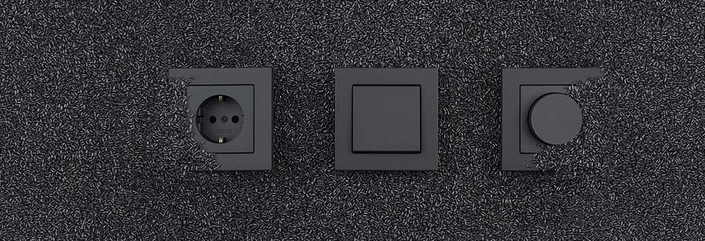 Das Bild zeigt eine schwarze Steckdose, einen Lichtschalter und einen Dimmer, die on schwarzem Kunststoffgranulat umgeben sind.