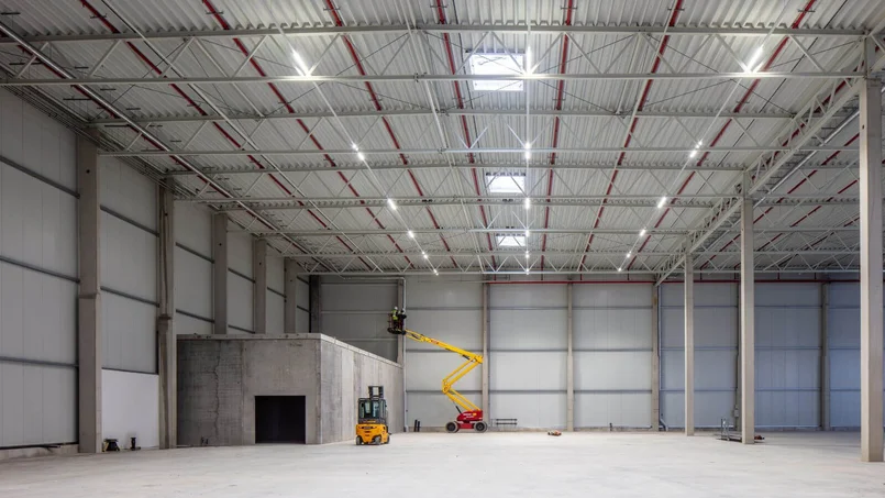 Eine große, leere Industriehalle mit hoher Decke; darin stehen ein Gabelstapler und eine Hebebühne, auf der zwei Personen an der Decke arbeiten.
