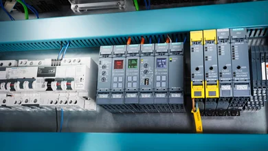 Zu sehen ist ein geöffneter Schaltschrank mit mehreren Siemens-Bausteinen, darunter Schutz-, Steuer- und Kommunikationsmodule. Rechts fallen die gelb markierten AS-i-Module auf, in der Mitte mehrere SIRIUS-Geräte mit Display und Tasten.