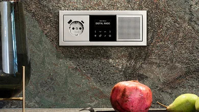 Flaches Radio zum Einbauen mit Steckdose direkt an der grauen Wand montiert. Davor liegen ein Granatapfel und eine Banane.